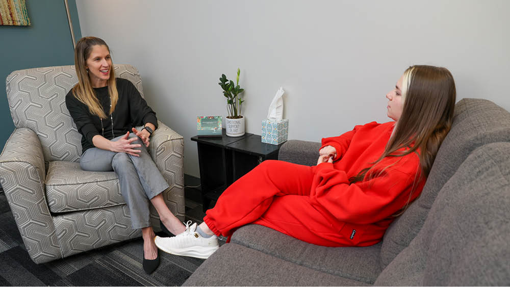 Heather Thomas, Grand View's Director of Counseling Services, in professional attire sits in an armchair, attentively listening to a student reclined on a sofa, conveying a supportive and calm atmosphere.
